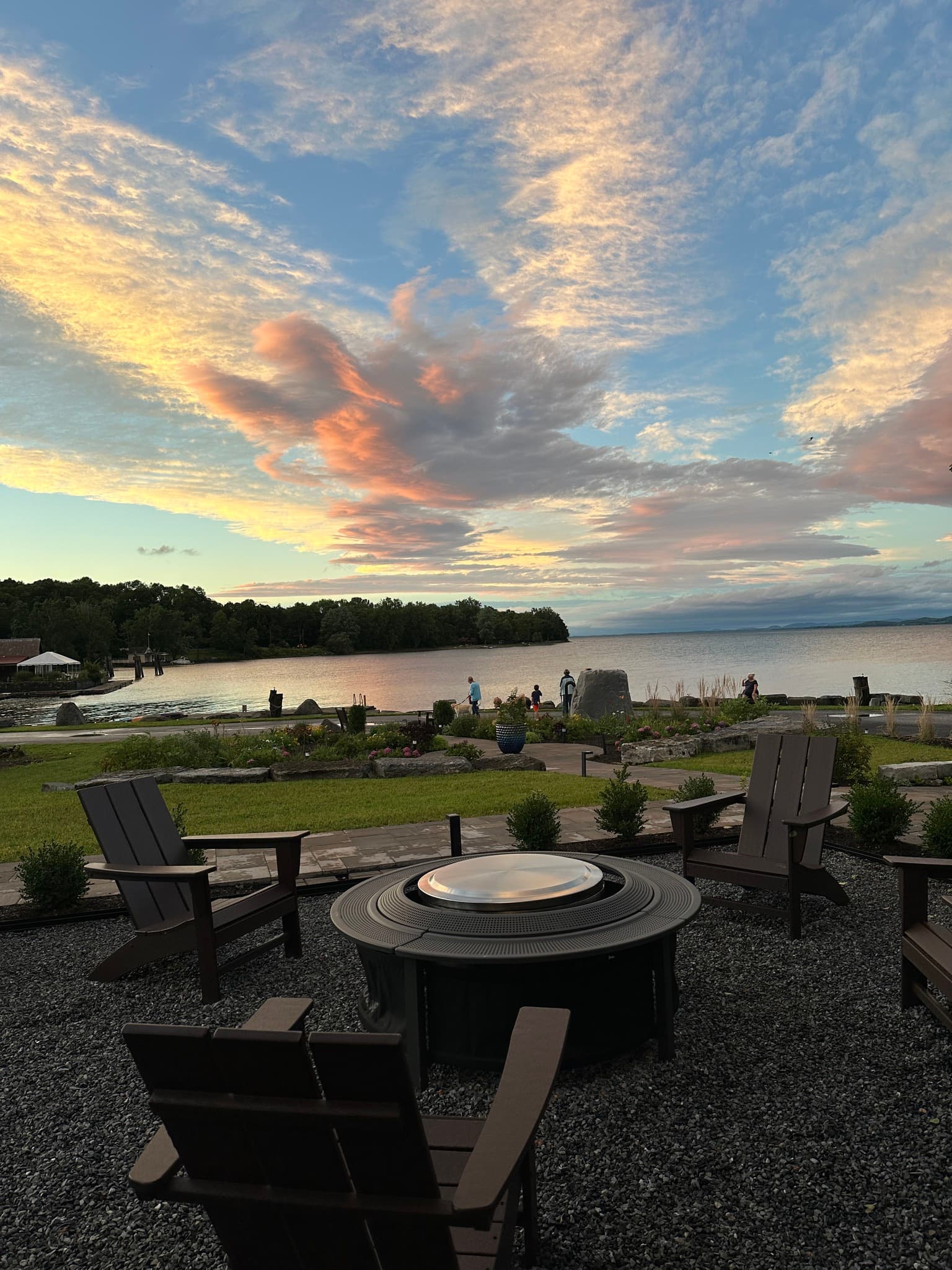 A peaceful lakeside view at sunset, featuring chairs around a fire pit and people enjoying the scenery.
