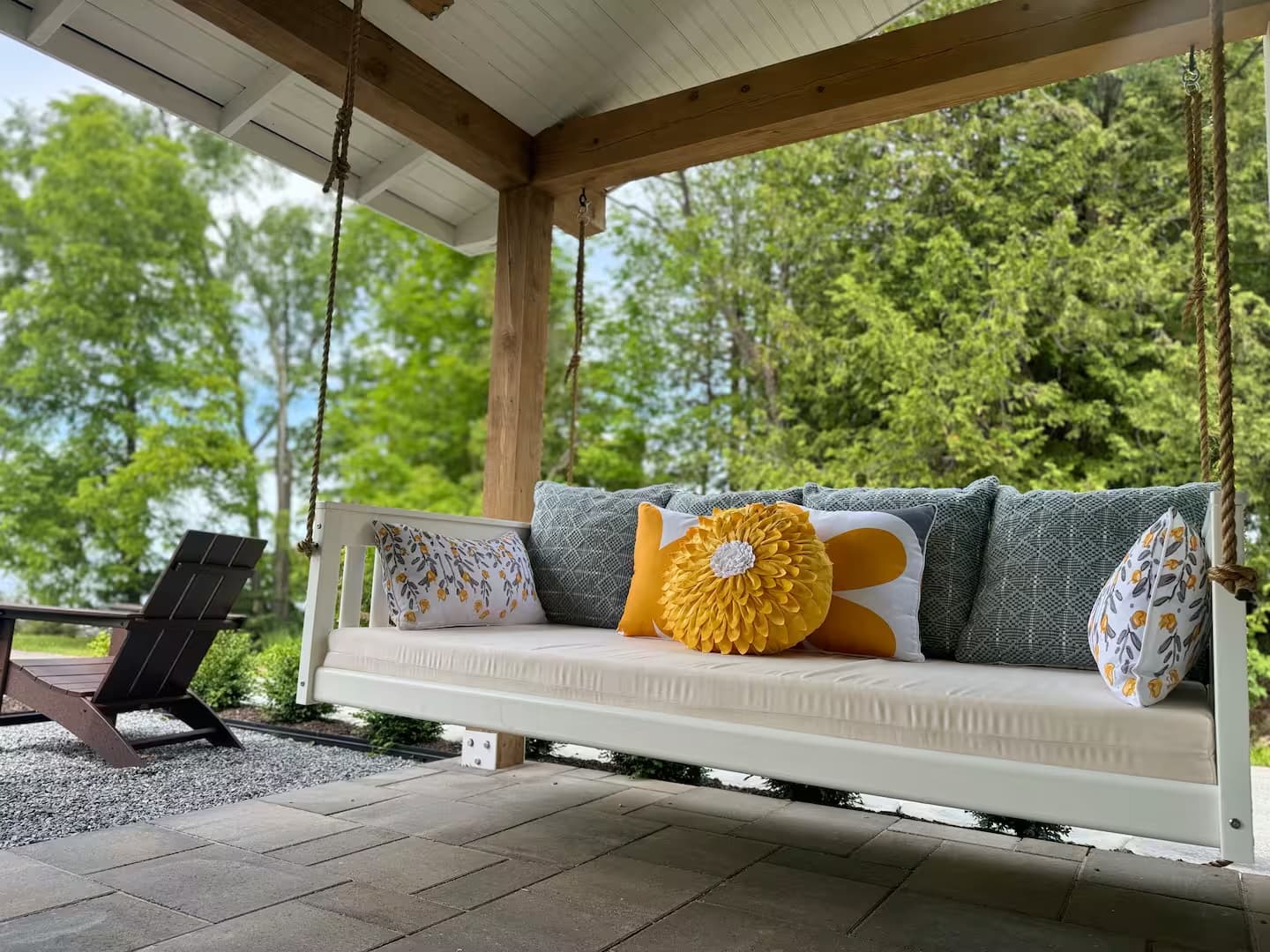 A serene porch swing with bright yellow and teal pillows sits on a patio. Lush green trees provide a beautiful, peaceful backdrop.