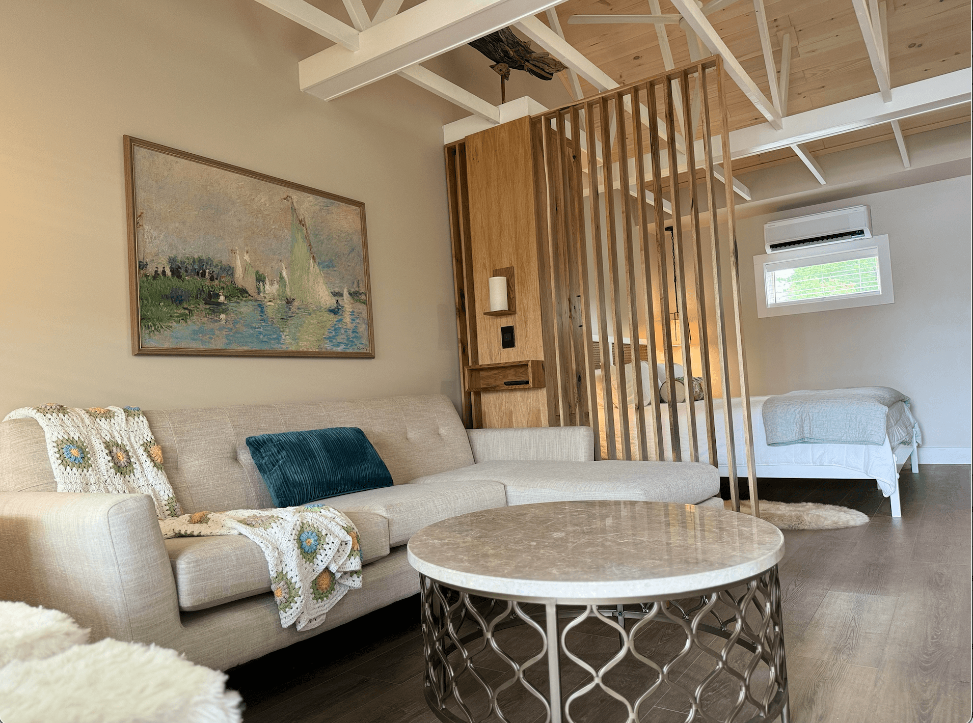 An inviting cottage interior features a cream sectional couch, a marble and metal coffee table, and a wood slat screen separating the living and sleeping areas.