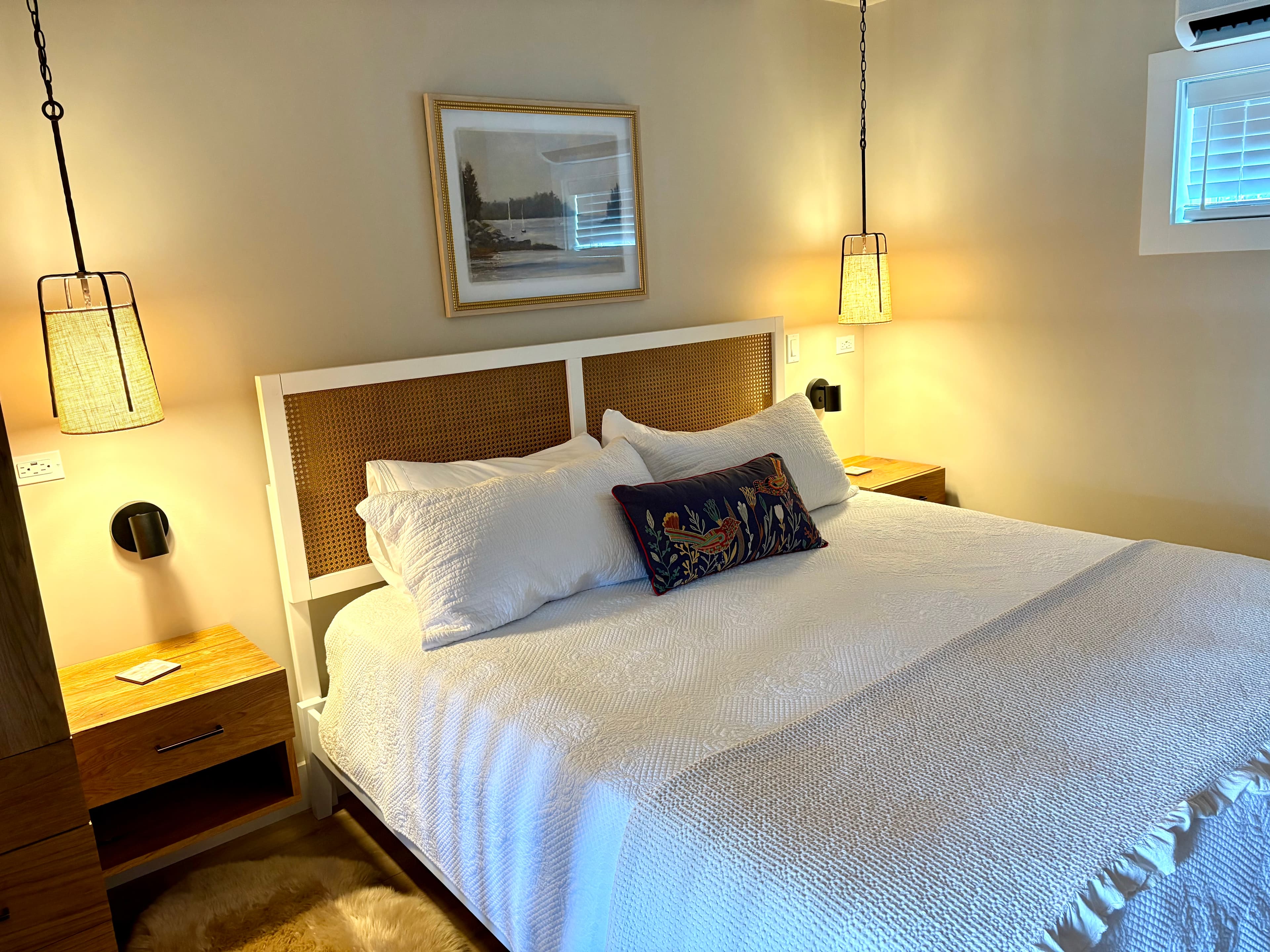 A cozy, well-lit bedroom features a king bed with white linens, a woven headboard, matching wood nightstands, and stylish hanging pendant lights.
