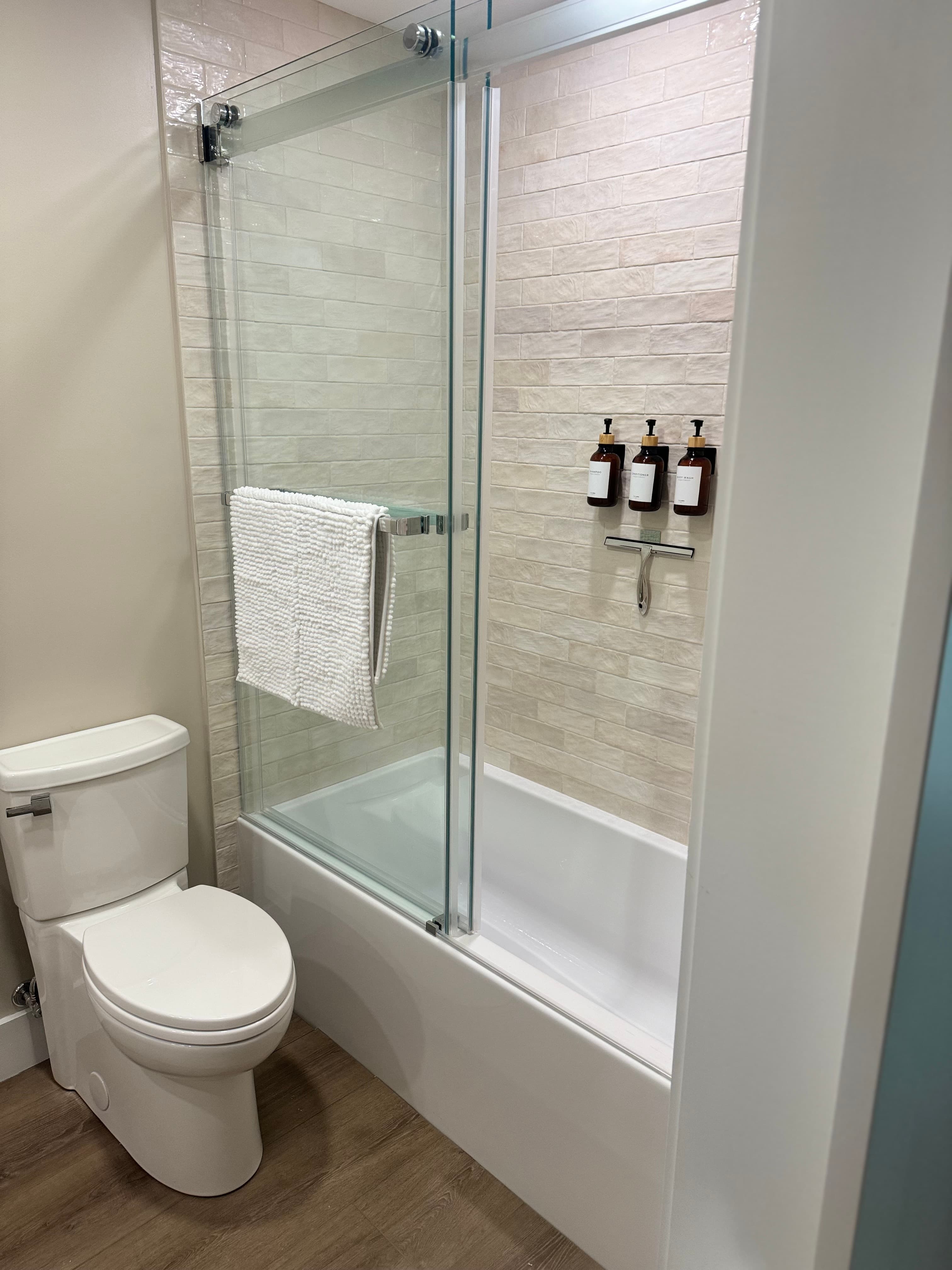 A modern bathroom with a white toilet and a soaking tub/shower combination. The shower features a glass door, tiles, and wall-mounted dispensers.