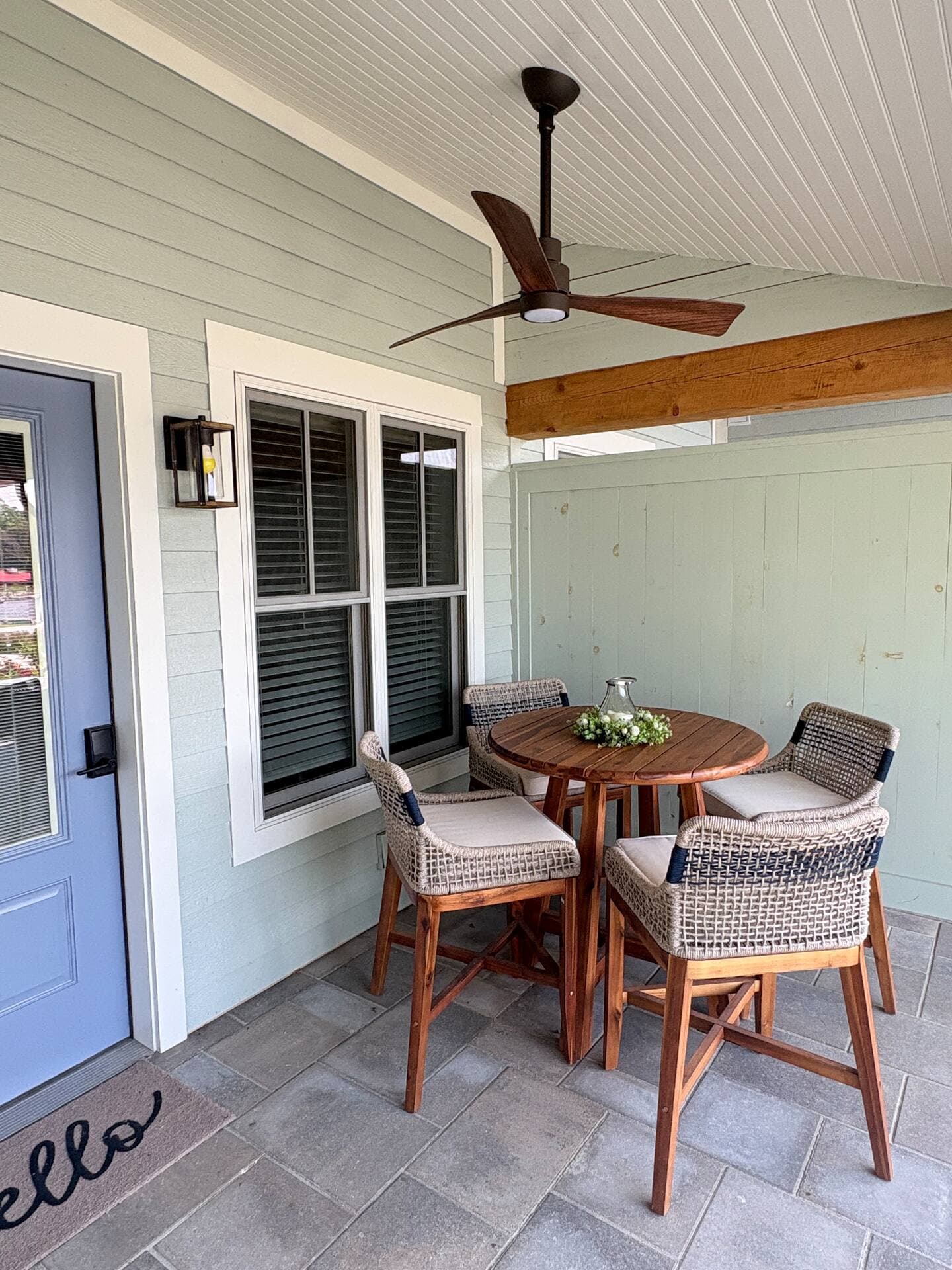 A welcoming porch with light blue siding features a small wood bar table and three counter-height woven chairs. A dark ceiling fan hangs above.