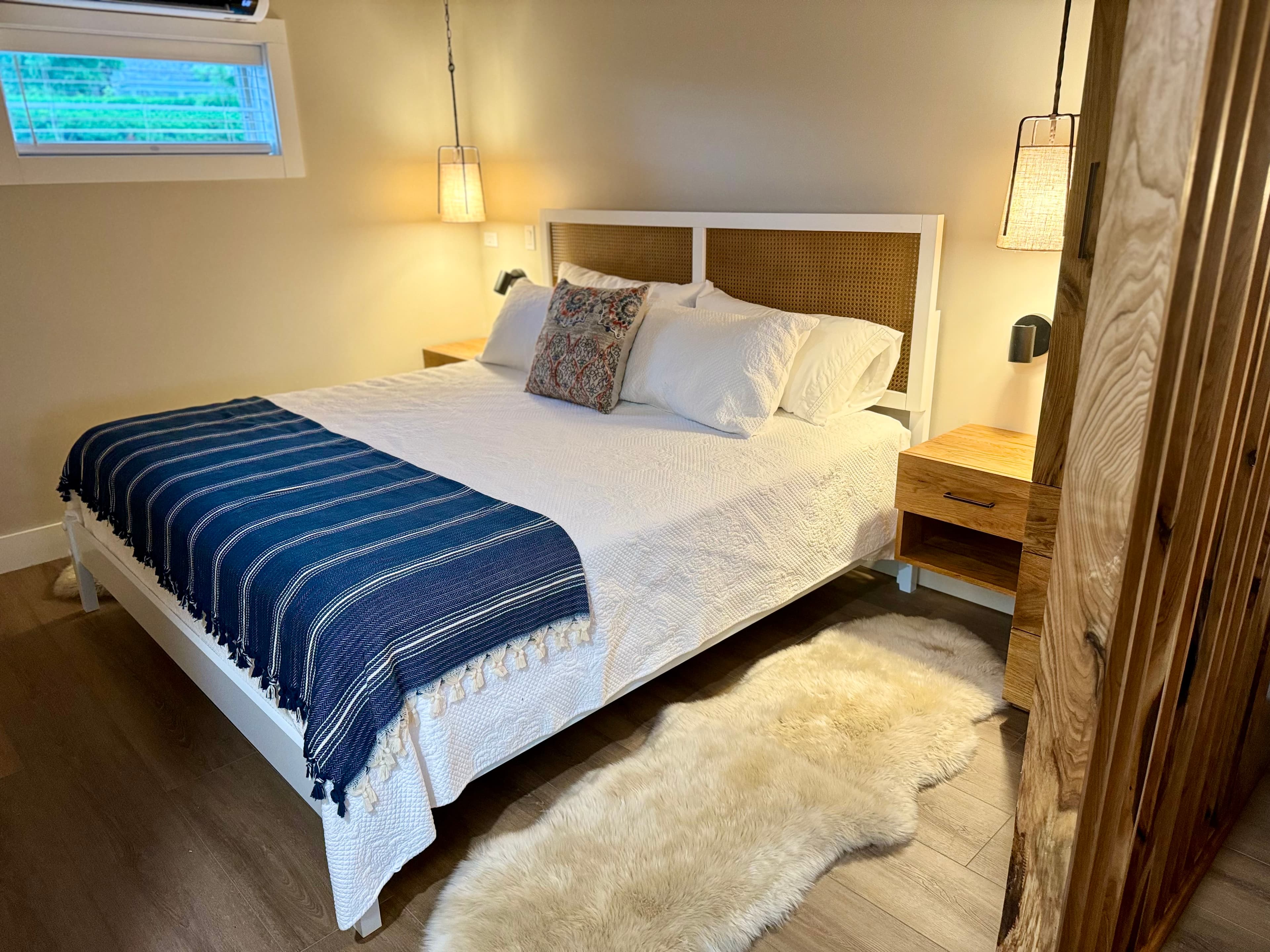 A bright and inviting bedroom features a king bed with a blue striped throw blanket, a woven headboard, and matching wood nightstands.
