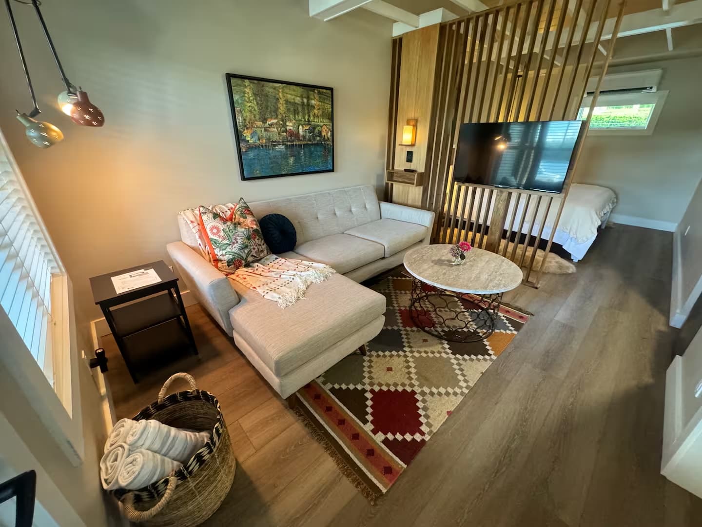 A cottage interior features a beige sectional couch, a colorful patterned rug, and a wood slat partition to separate the living and sleeping areas.