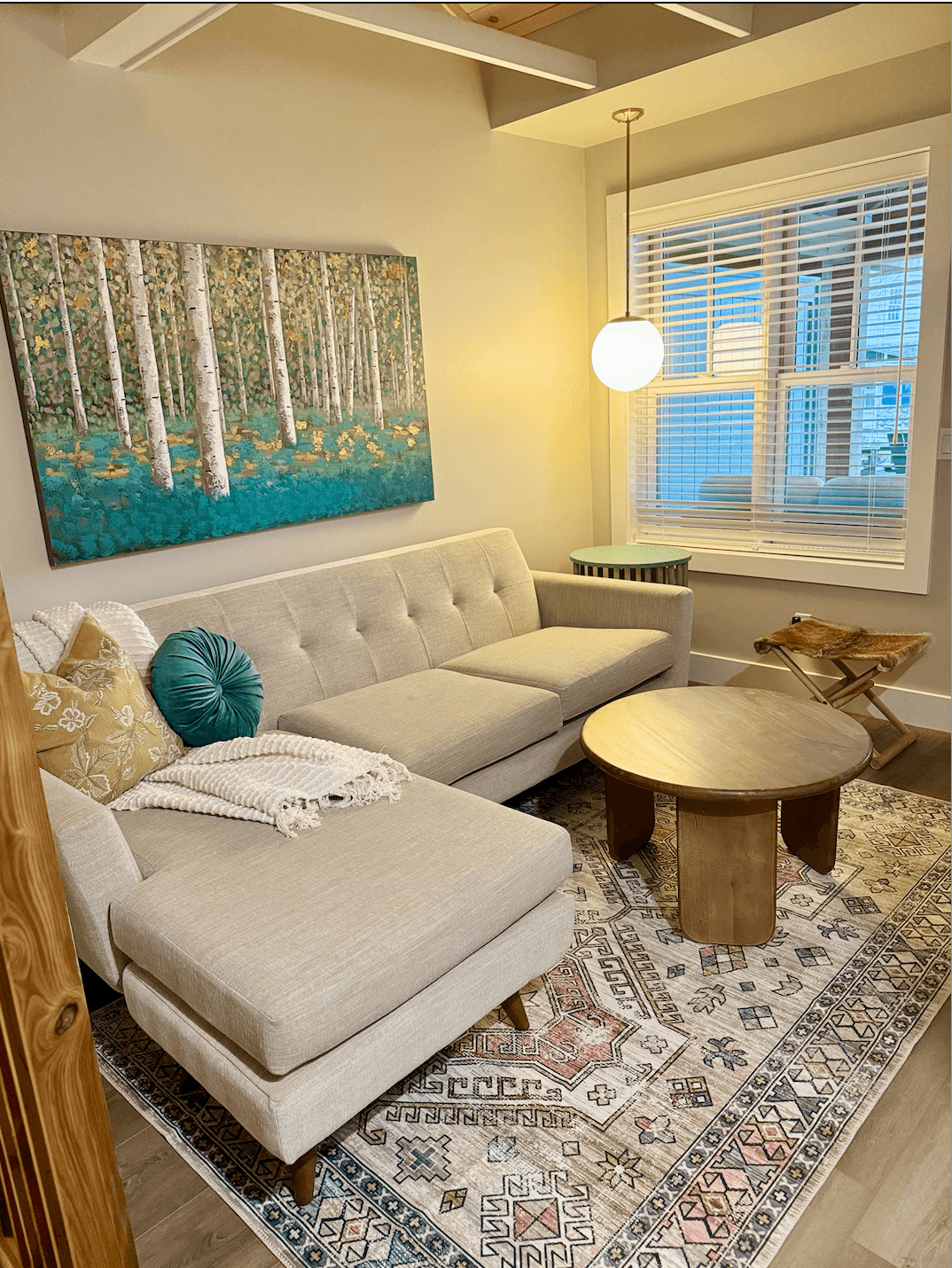 A stylish living area features a beige chaise lounge sofa, a round wood coffee table, and vibrant birch tree artwork over the couch.