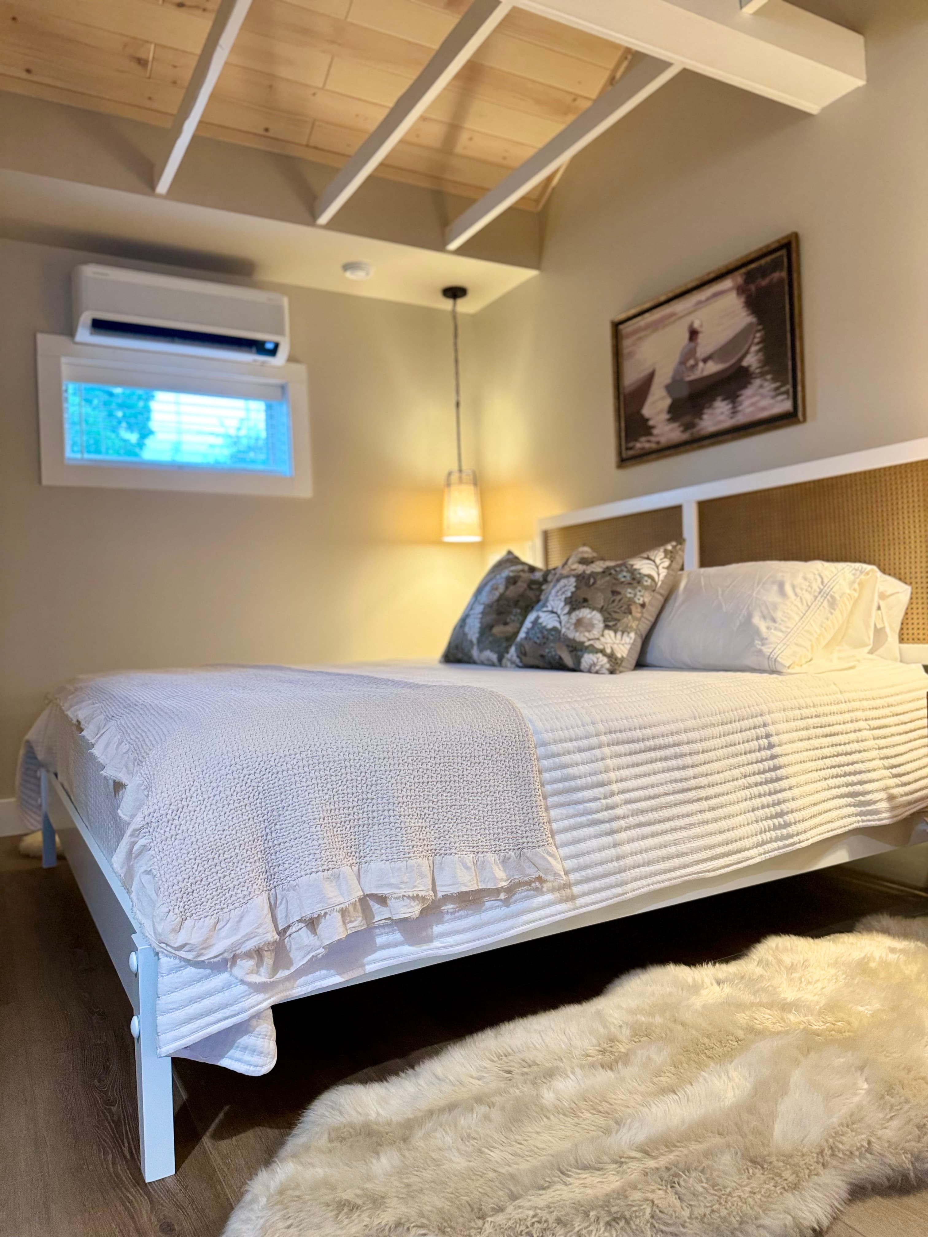A bright and cozy bedroom features a king bed with white linens, a woven headboard, wood exposed beams on the ceiling, and a furry rug.