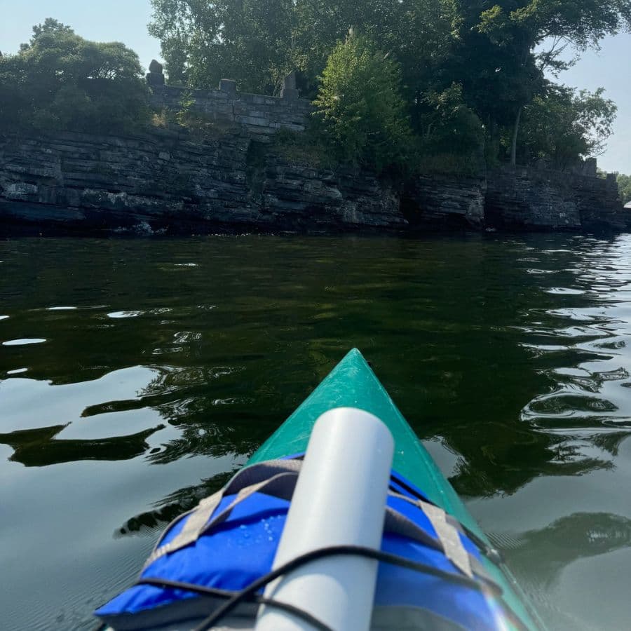 A kayak is seen navigating calm waters near a rocky shoreline lined with foliage.