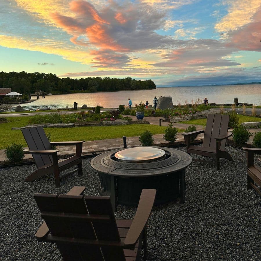 A serene lakeside scene at sunset, featuring a fire pit surrounded by wooden chairs and people enjoying the beautiful view.