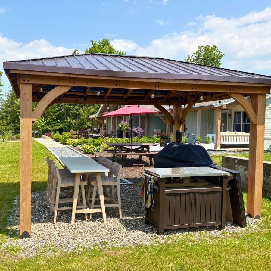 A wooden gazebo with a table, chairs, and a grill, set in a landscaped yard under a blue sky.