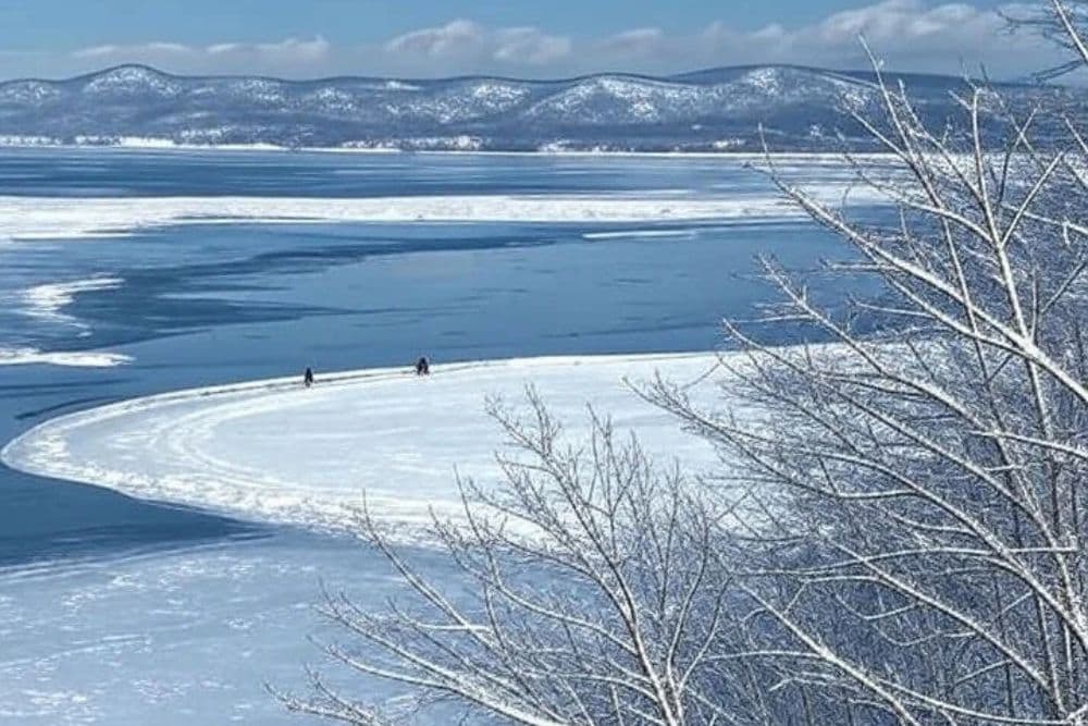 A serene winter landscape featuring two people on a snow-covered lake surrounded by mountains and blue skies.